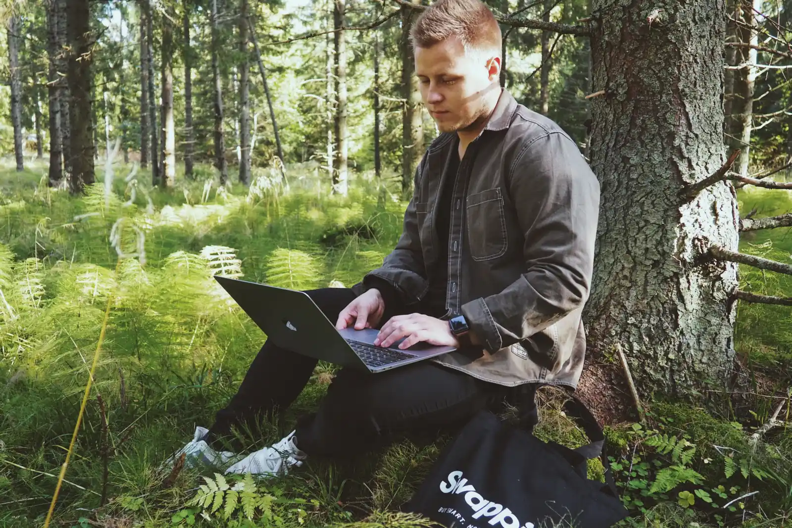 Man typing on a laptop while sitting outdoors in a green forest