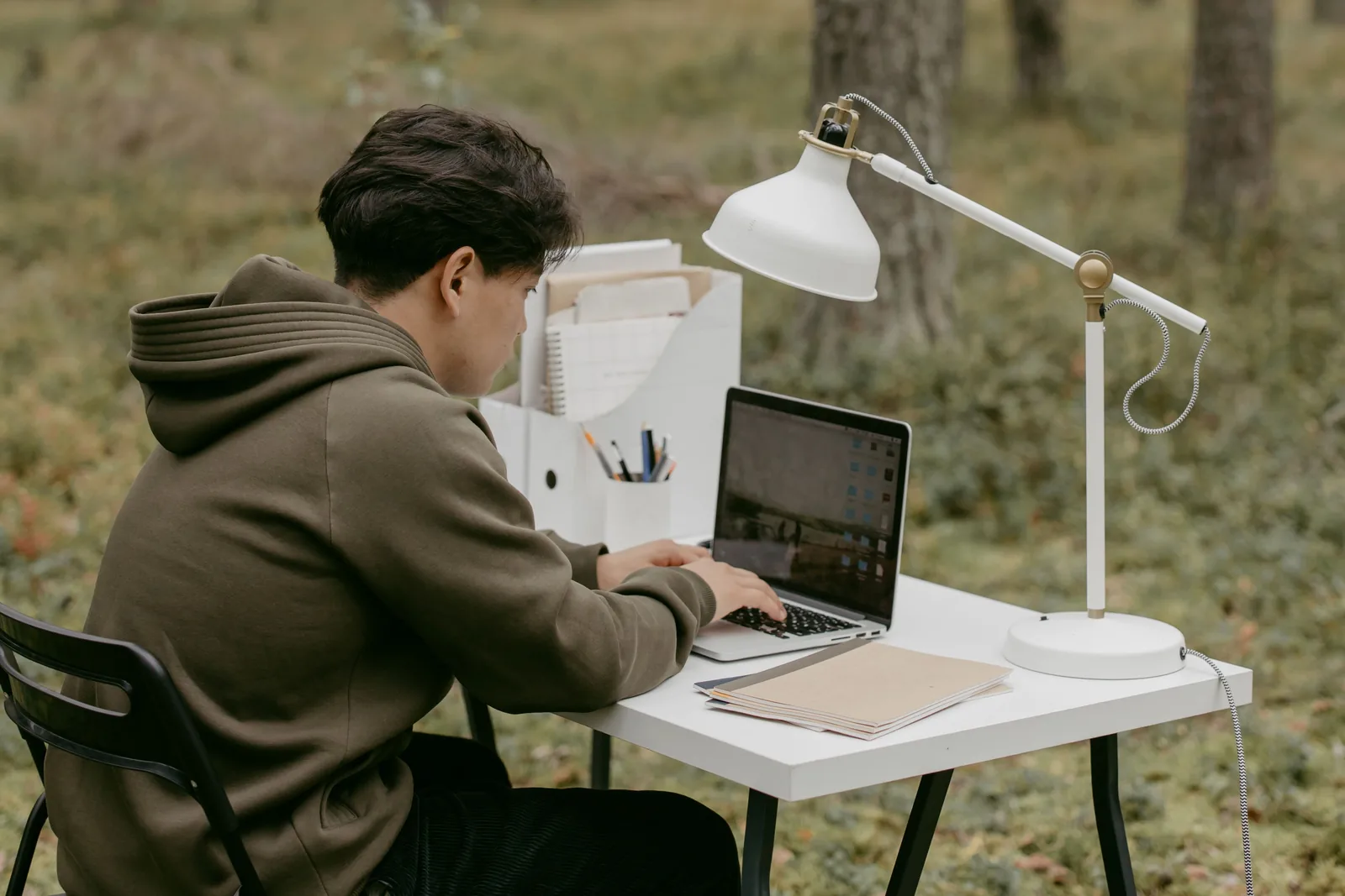 Man in a hoodie working on a laptop at an outdoor desk among trees