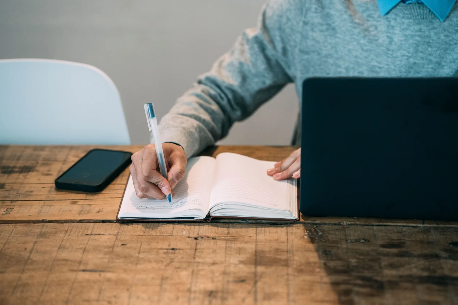 Person writing in a planner next to a laptop on a wooden desk